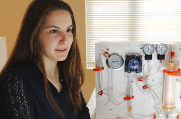 A hospital-grade dialysis machine in soft clinical light — control screen, peristaltic pump head, arterial and venous ports — the kind of $20,000–$80,000 equipment a 15-year-old's $1,000 alternative is built to bypass for the 2.3 million patients who cannot reach one