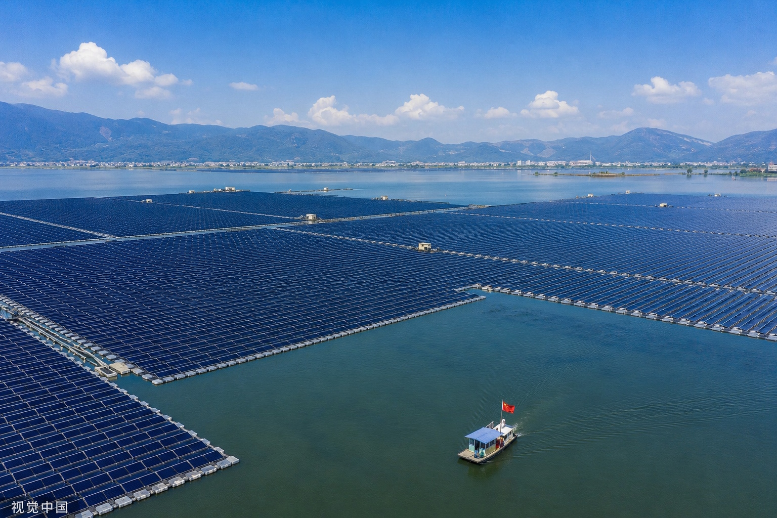 Rows of solar photovoltaic panels mounted on floating platforms across a calm lake surface, reflecting blue sky and creating a mosaic of energy generation on water